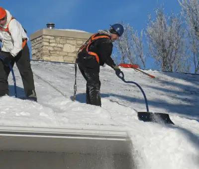 Professional technicians performing roof snow removal with safety harnesses and shovels in Dover, DE